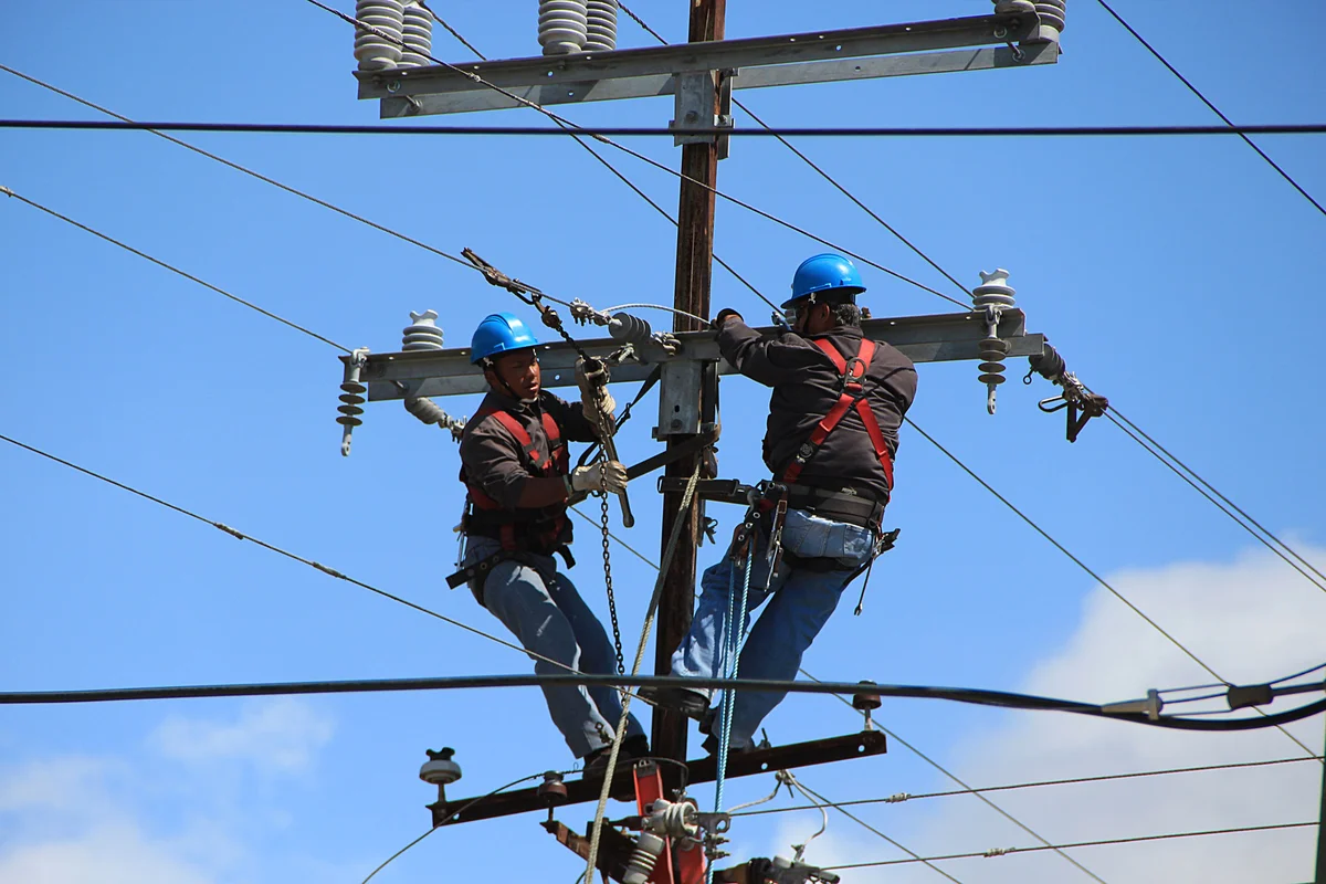 Two linemen in hard hats and fall protection harnesses working crossarm construction on a wood pole